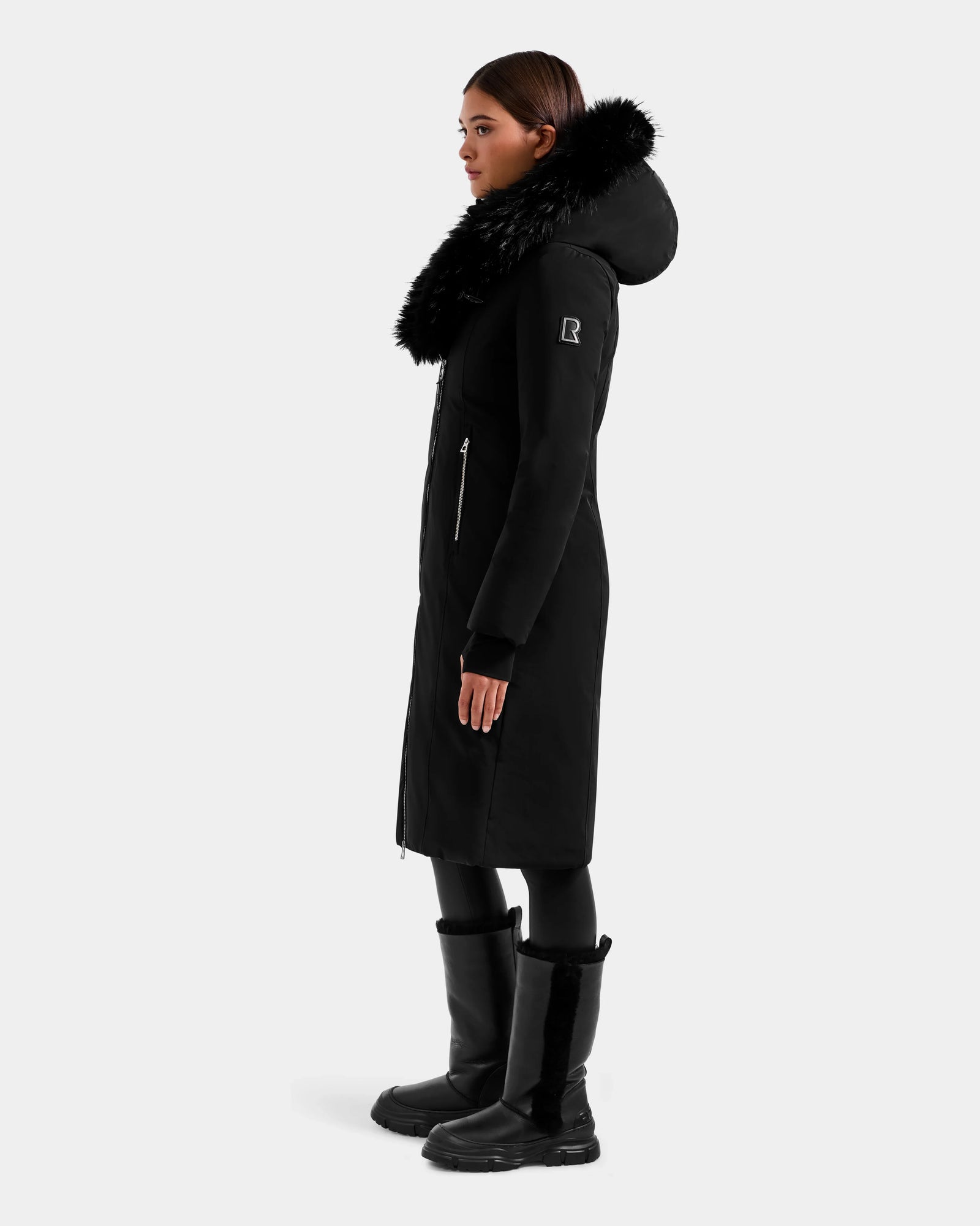 A woman in profile models the Rudsak JANIS - L125558 Black winter coat with a large faux fur hood, paired with black leggings and tall black boots, set against a plain white background.