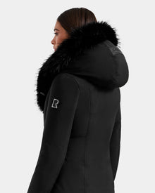 Woman wearing the Rudsak JANIS - L125558 Black down parka, seen from the side and back. The coat has a large faux fur hood, patch logo on the sleeve, and is set against a plain light gray background.