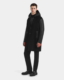 A man stands against a plain background wearing the Rudsak JESSE - M125504 Black men’s wool coat with leather sleeves, paired with black pants, black shoes, and a dark hoodie. He looks forward with a neutral expression.
