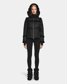 A woman stands facing forward against a plain white background wearing the Rudsak JULIANA - L124509 Black/Black women's short down puffer with a hood, black leggings, and black winter boots.