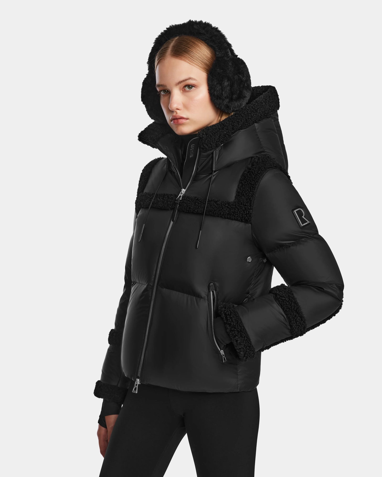A woman wears the Rudsak JULIANA - L124509 Black/Black RDS-certified down jacket with fur trim and matching earmuffs, paired with black leggings, standing against a white background and looking slightly to the side.