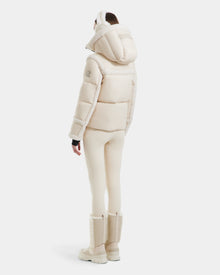 A person faces away, displaying Rudsak’s JULIANA - L124509 Vanilla women’s short down puffer hooded jacket in light cream, paired with matching pants, earmuffs, and fur-trimmed boots, set against a plain white background.