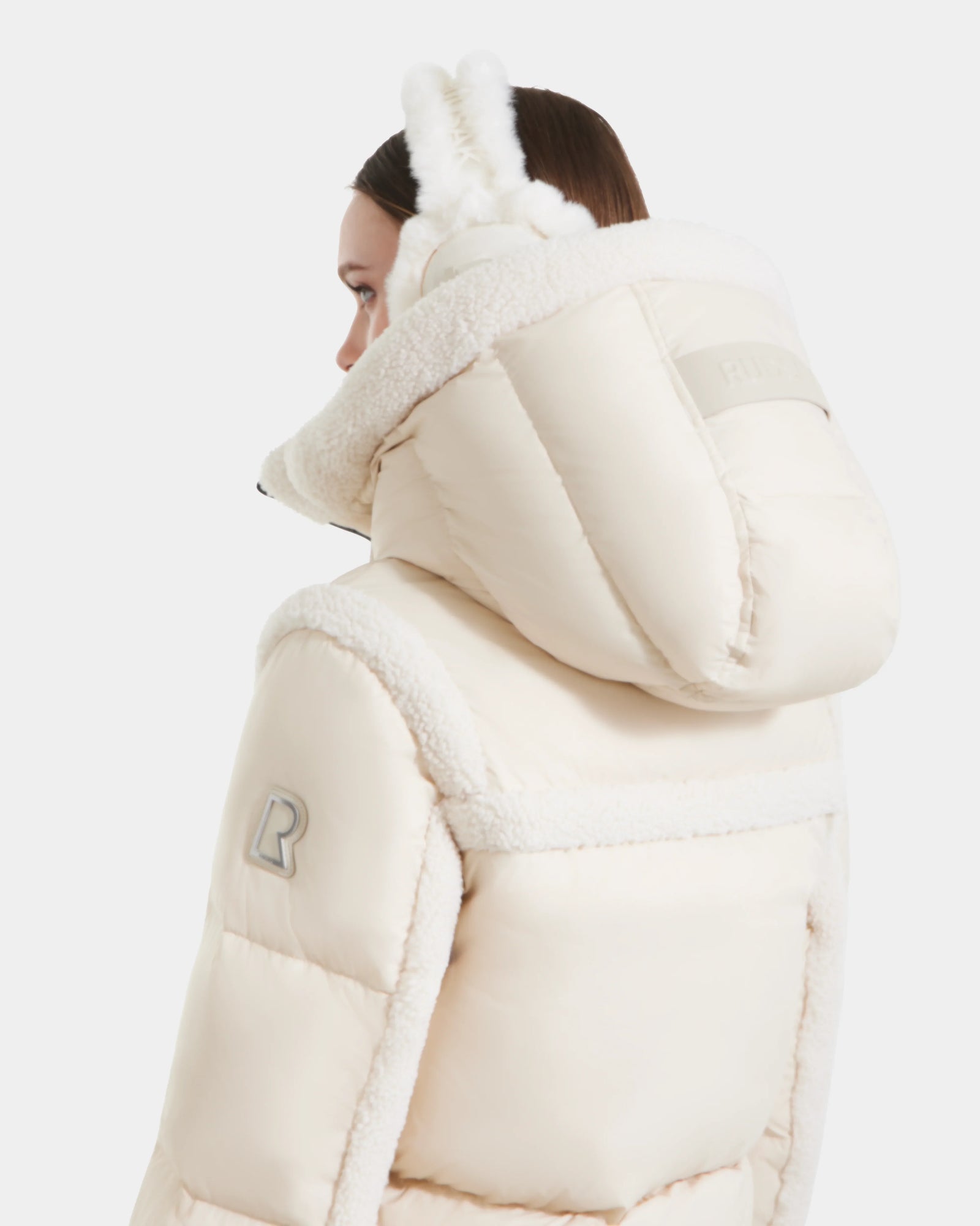 Wearing the Rudsak JULIANA - L124509 Vanilla women’s short down puffer with a large hood and shearling trim, a person holds fluffy earmuffs to their head, seen from the back and side against a white background.