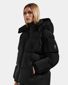 A woman with straight blonde hair wears the Rudsak KARLEE M - 8125681 Black women’s down puffer jacket with a high collar and hood, hands in pockets, standing against a light gray background—a stylish winter choice.