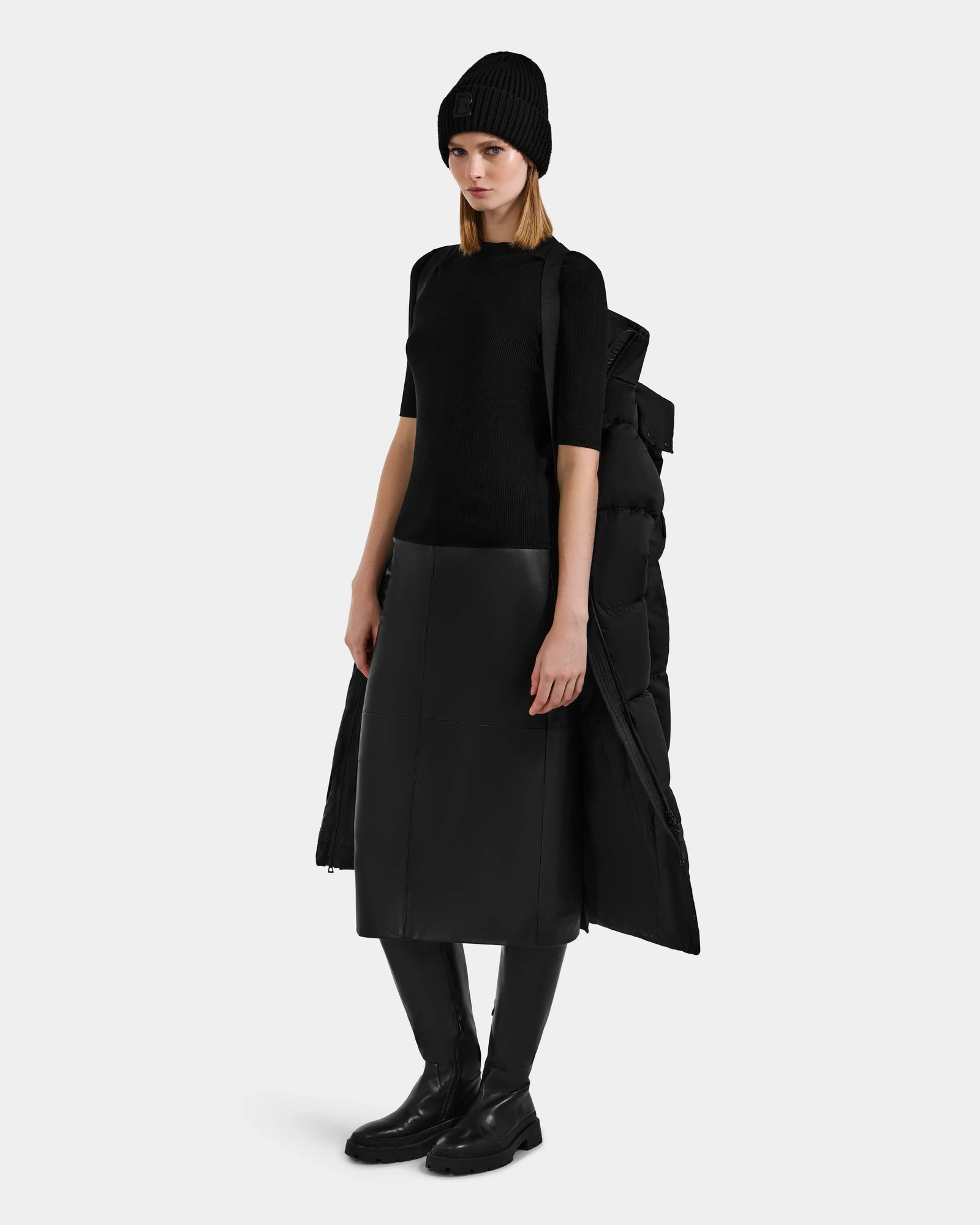 A woman stands against a plain background wearing the Rudsak KARLEE XL - 8125683 Black women’s maxi down puffer jacket draped over her shoulders—a true winter essential for extreme cold-weather performance.