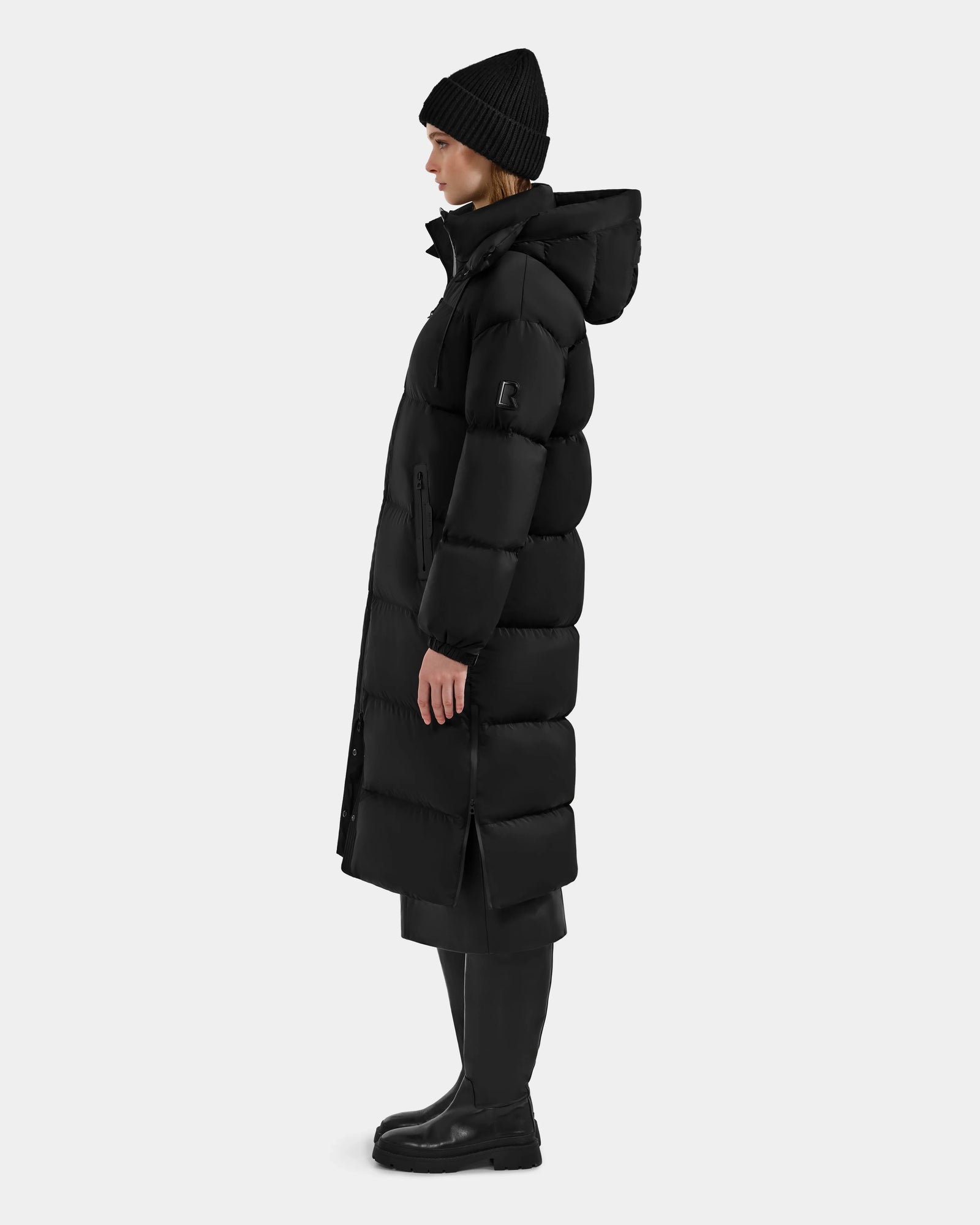 Side view of a person wearing the Rudsak KARLEE XL - 8125683 Black women’s long maxi down puffer jacket, styled with a black beanie, pants, and boots against a plain light background—perfect for winter warmth.
