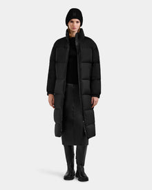 A person wearing the Rudsak KARLEE XL - 8125683 Black women’s long maxi down puffer jacket, styled with black accessories, stands facing forward against a plain gray background—a true essential for extreme winter cold.
