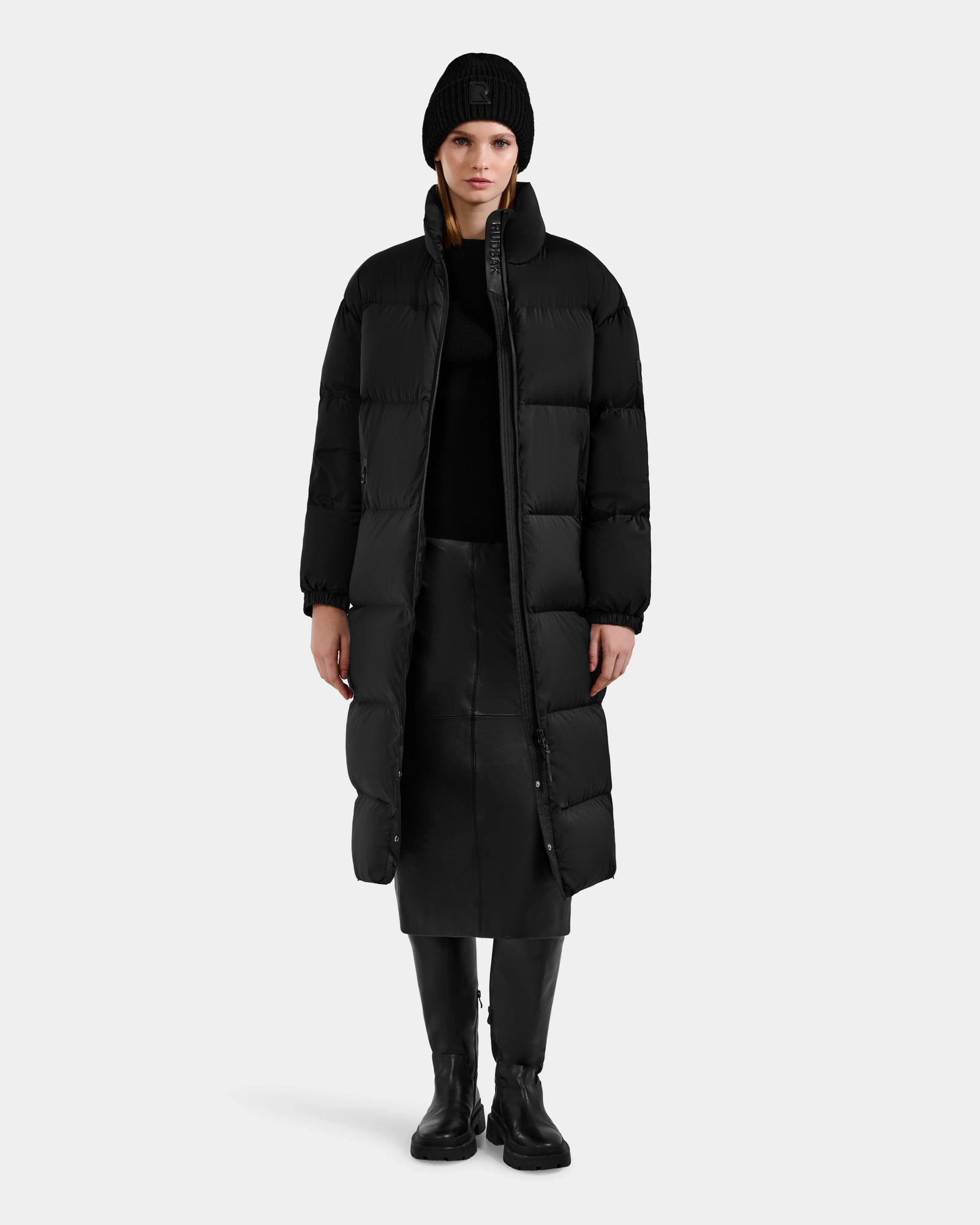 A person wearing the Rudsak KARLEE XL - 8125683 Black women’s long maxi down puffer jacket, styled with black accessories, stands facing forward against a plain gray background—a true essential for extreme winter cold.