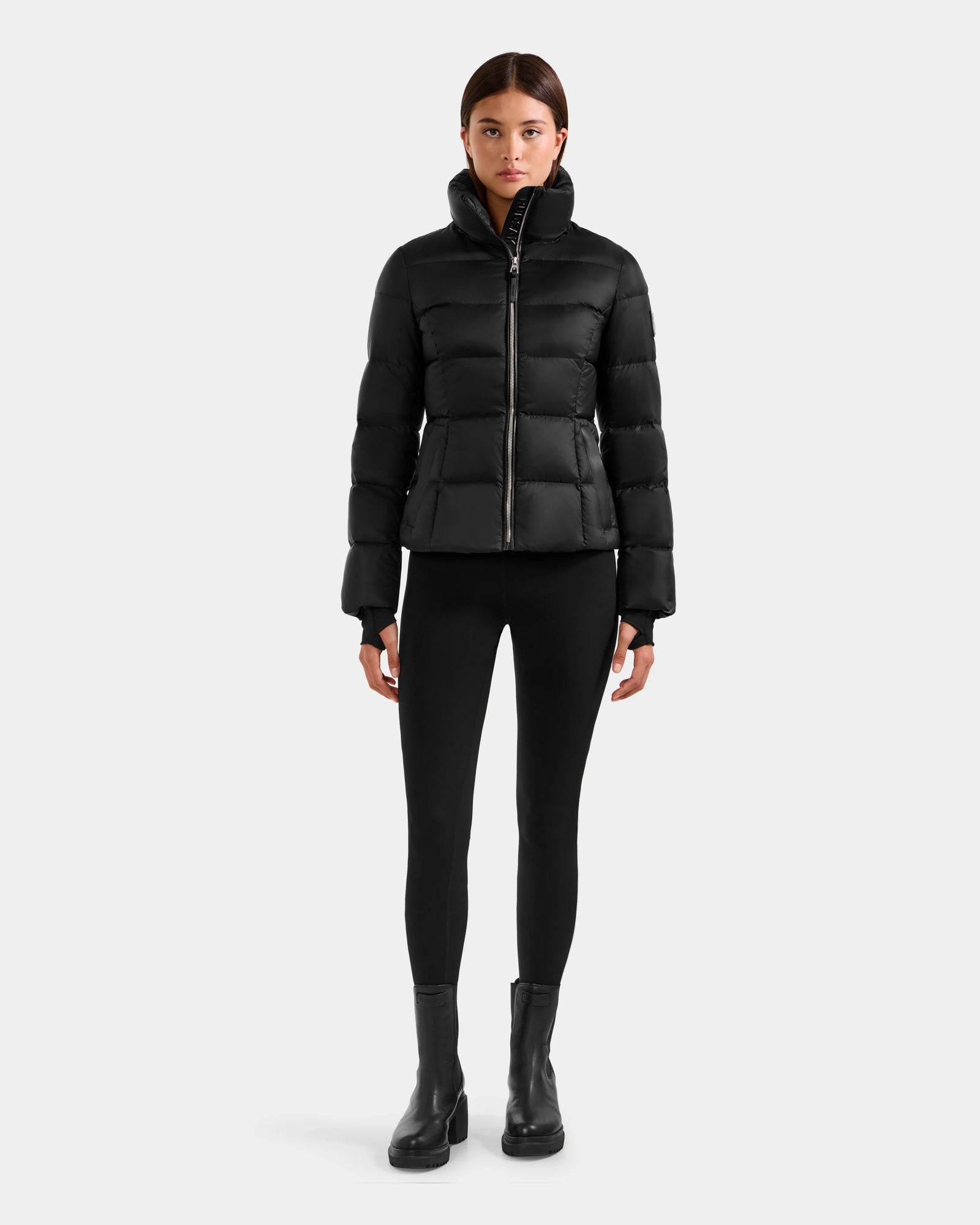 A woman faces forward wearing the Rudsak KATIA - L125564 Black puffer jacket with RDS-certified down, paired with black leggings and ankle boots, set against a plain light gray background.