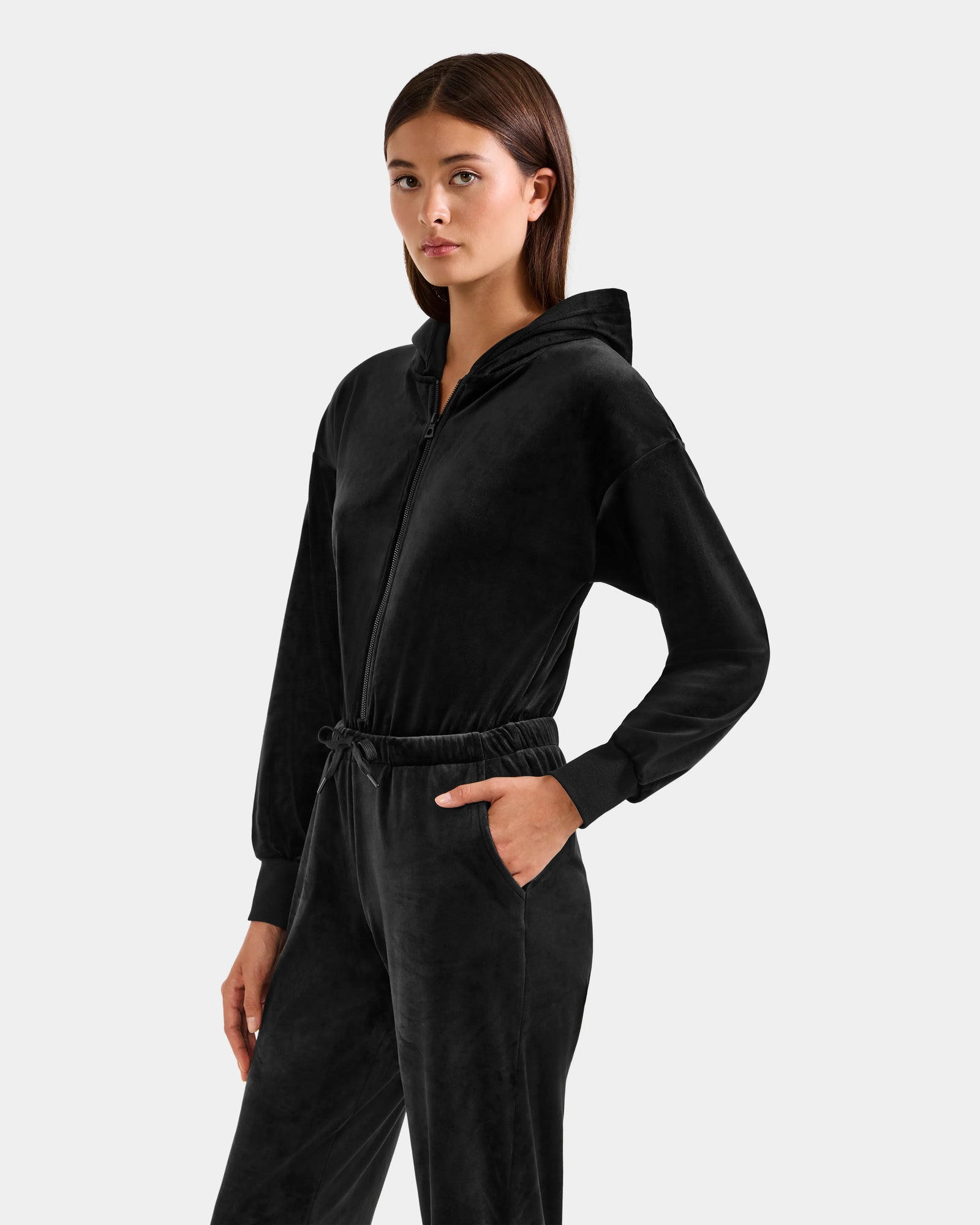 A woman with straight brown hair models the Rudsak KETZY - L525503 Black velour hooded jumper, featuring long sleeves, side pockets, and a drawstring waist, against a plain light gray background—modern women's loungewear.