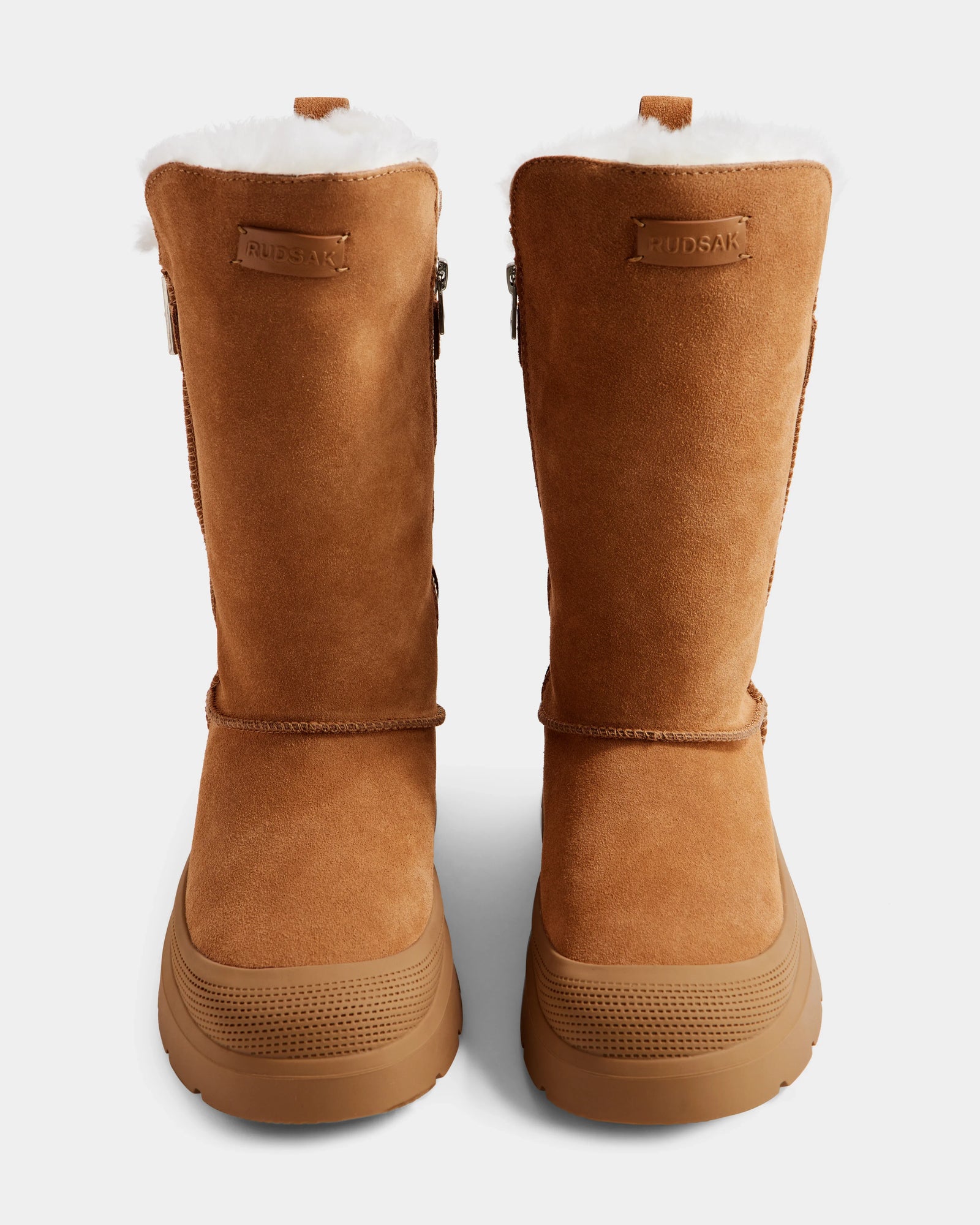 The KLAIR - L225027 Tan by Rudsak are women’s tan suede winter boots with white wool lining, rubber soles, and a leather patch near the top embossed with the Rudsak brand name, shown from the front.