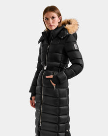 A woman wears the Rudsak LILITH L - L125528 Black long puffer coat with a tan faux fur-trimmed hood, standing against a plain light background and looking directly at the camera.