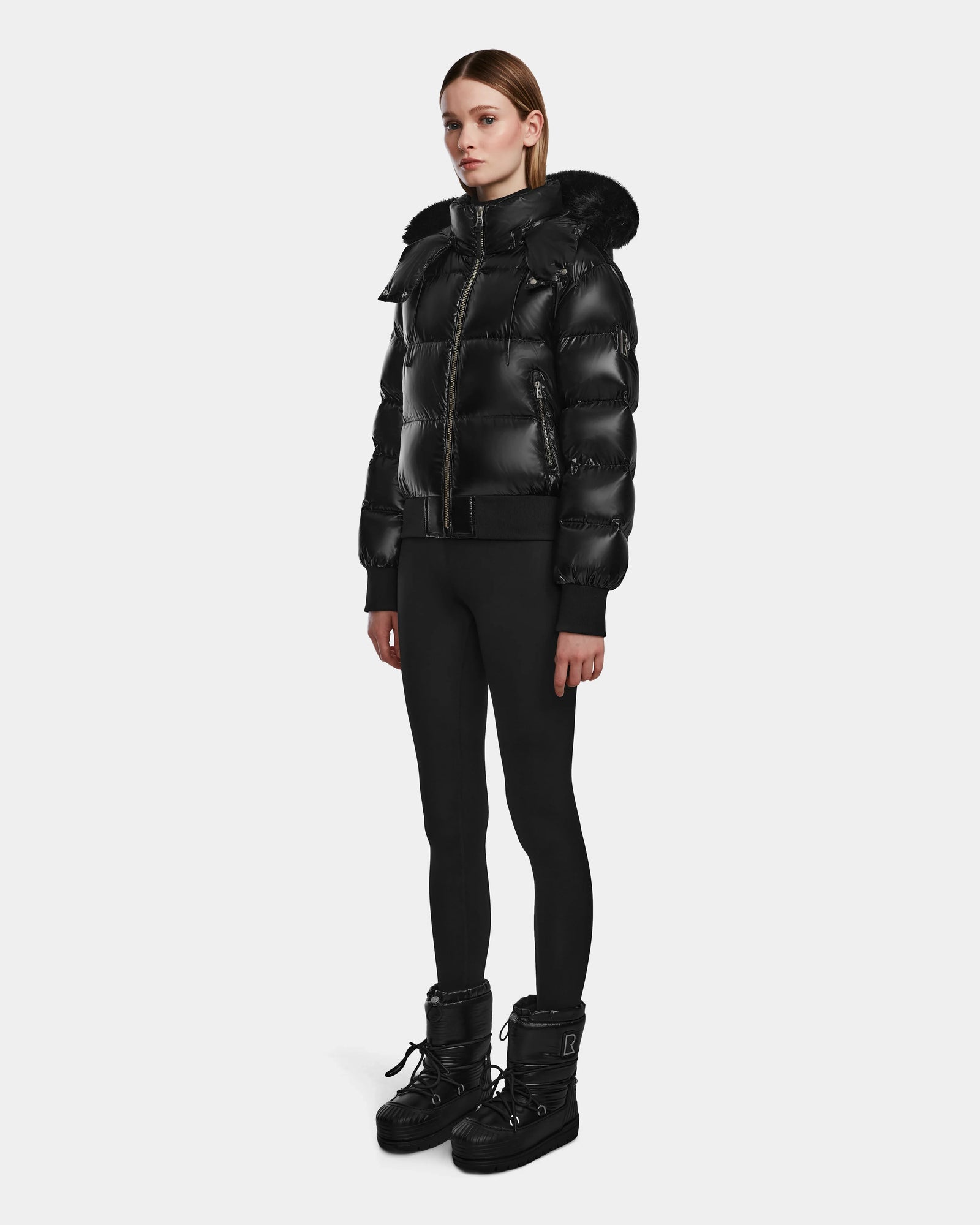 A woman faces forward against a white background, wearing the Rudsak MAUDE Q-S - L124553 Black quilted down bomber with a fur-trimmed hood, paired with black leggings and winter boots.