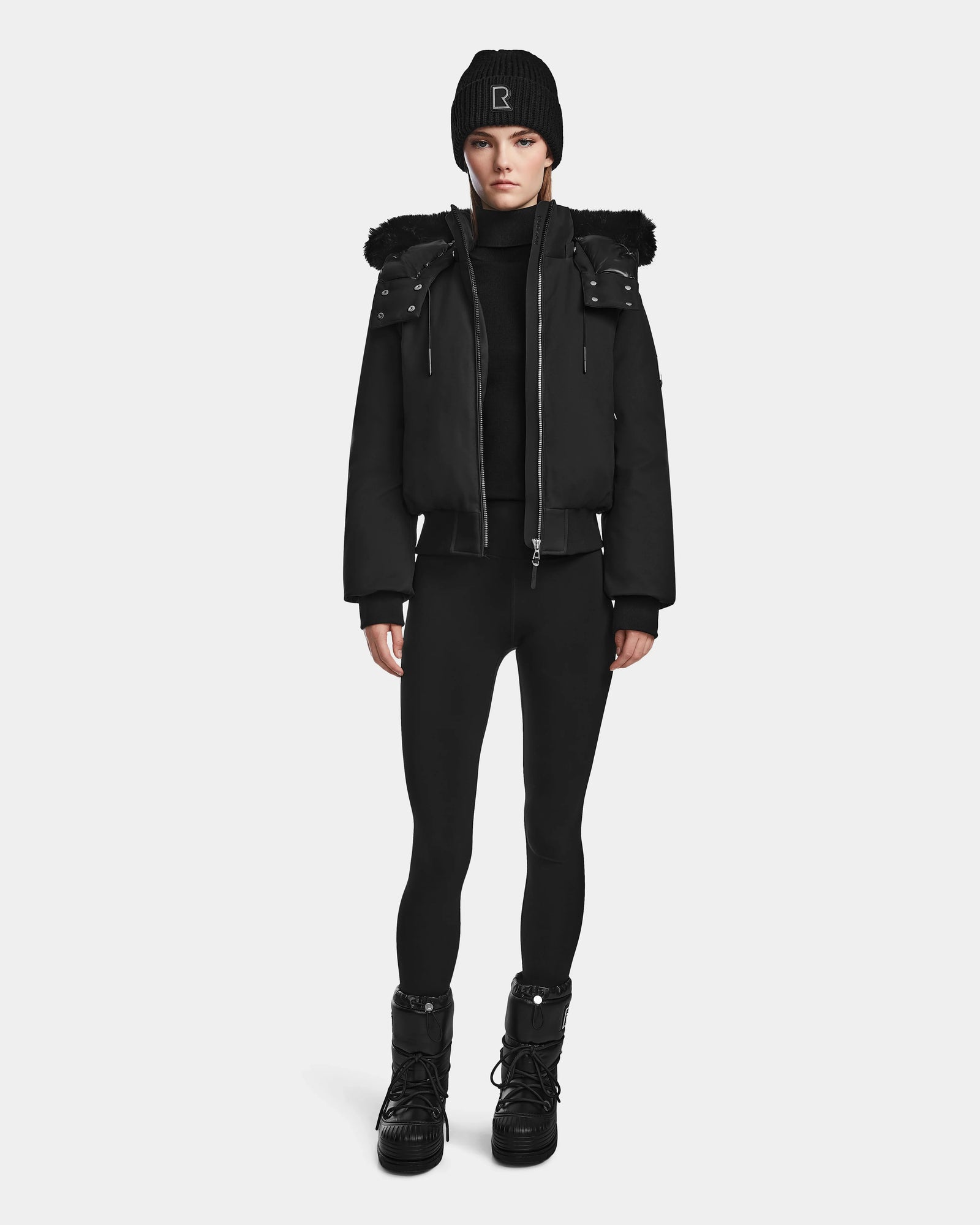 A person stands facing forward in a black Rudsak MAUDE - L124522 jacket with fur trim, paired with a black beanie, turtleneck, leggings, and lace-up boots against a plain white background.