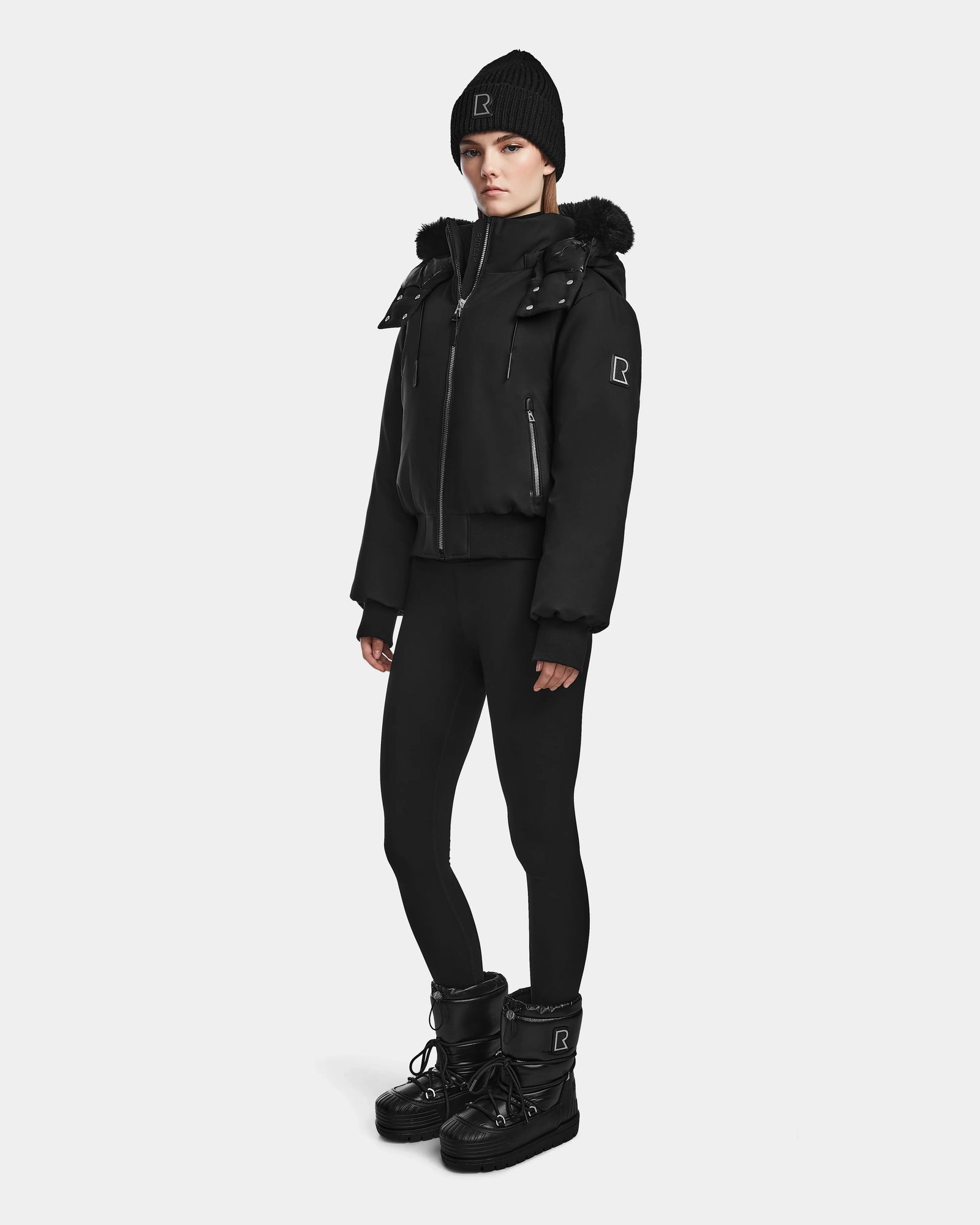A person faces forward wearing the Rudsak MAUDE - L124522 Black/Black winter jacket with a fur-trimmed hood, styled with a black beanie, leggings, and boots against a plain white background.