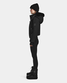 A person in profile wears the Rudsak MAUDE - L124522 Black/Black women's down bomber, paired with black leggings and black boots, standing against a plain white background.