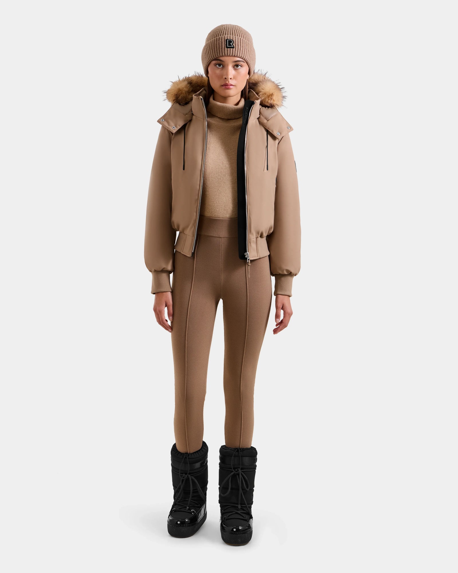 A person faces forward against a plain white background, wearing the Rudsak MAUDE - L124522 Sand winter jacket, paired with a matching turtleneck, fitted tan pants, tan beanie, and black winter boots.