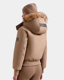A person models the Rudsak MAUDE - L124522 Sand jacket, featuring premium down insulation, a fur-trimmed hood, and a black patch, paired with a matching knit beanie and fitted pants, standing against a white background.