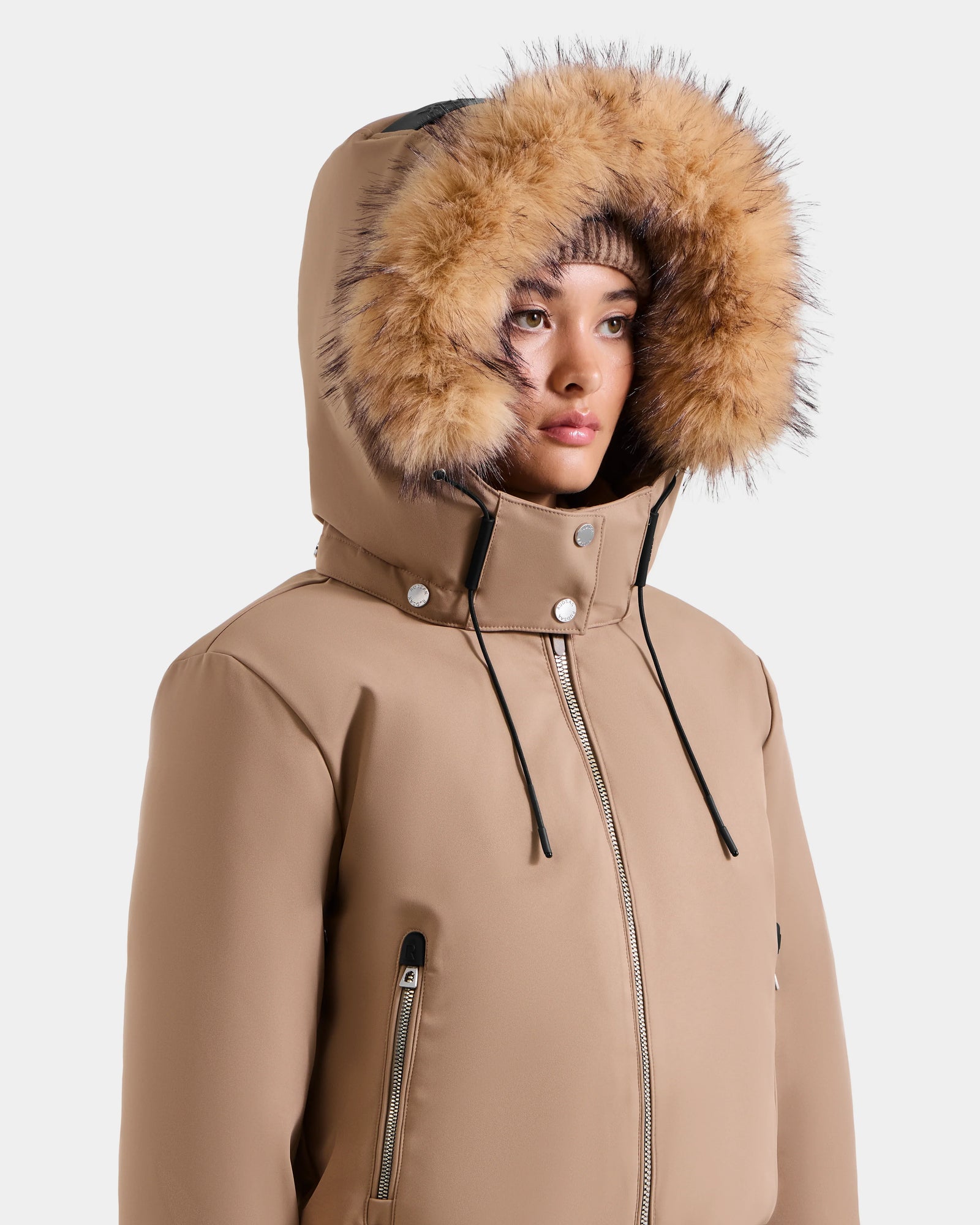 A person wears the Rudsak MAUDE - L124522 Sand jacket, tan with a fur-lined hood up, black drawstrings, silver zippers, snap buttons, and premium down insulation. They look slightly to the side against a plain white background.