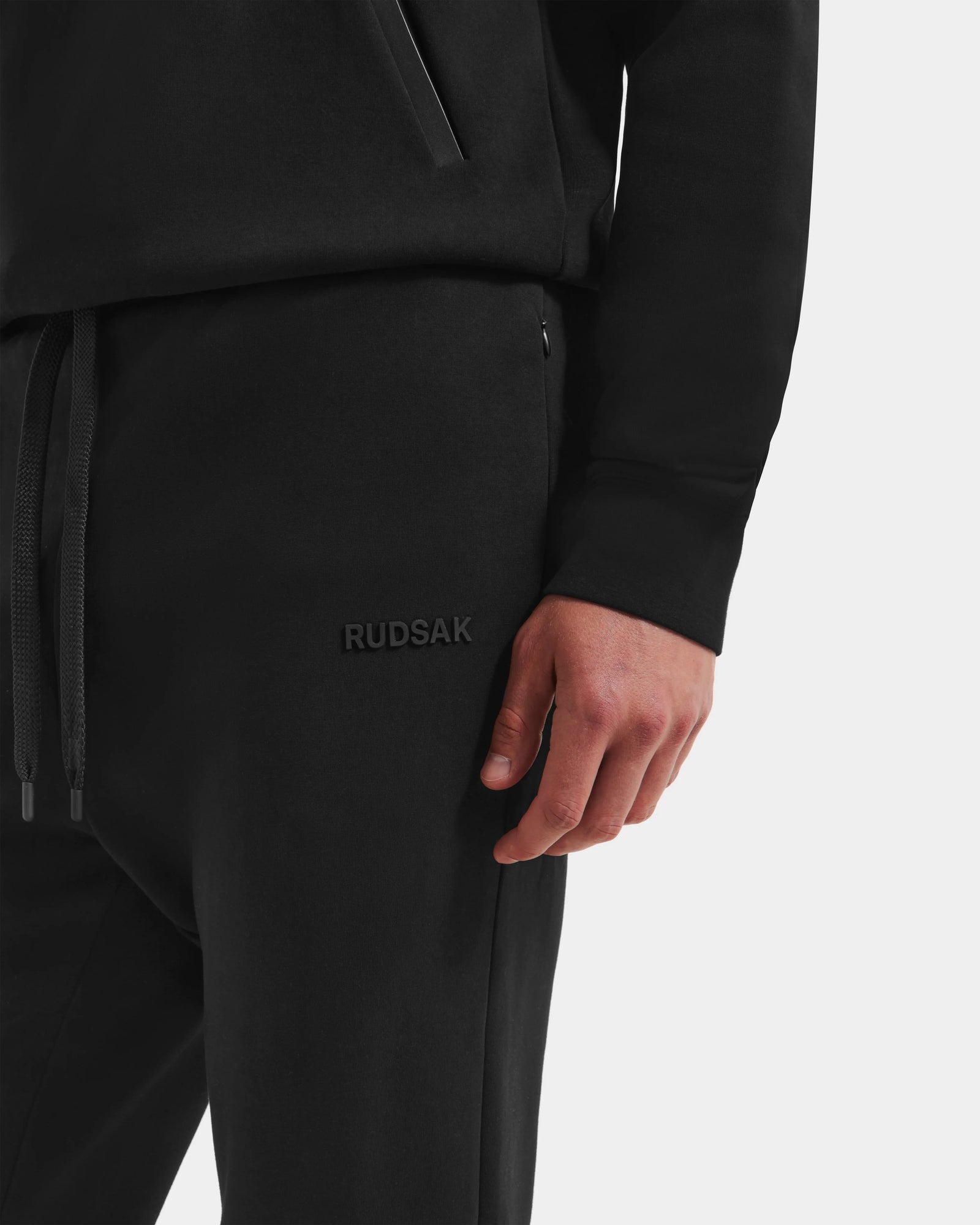A person showcases the Rudsak MAXTON - U525007 Black M jogger, featuring "RUDSAK" embroidery. Only the lower torso and right hand are visible, emphasizing this unisex cotton-blend pant designed for everyday comfort.