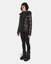 A woman faces forward in a Rudsak MIRRY S - L123545 Black quilted down coat, styled with black leggings, winter boots, and earmuffs against a plain white background.