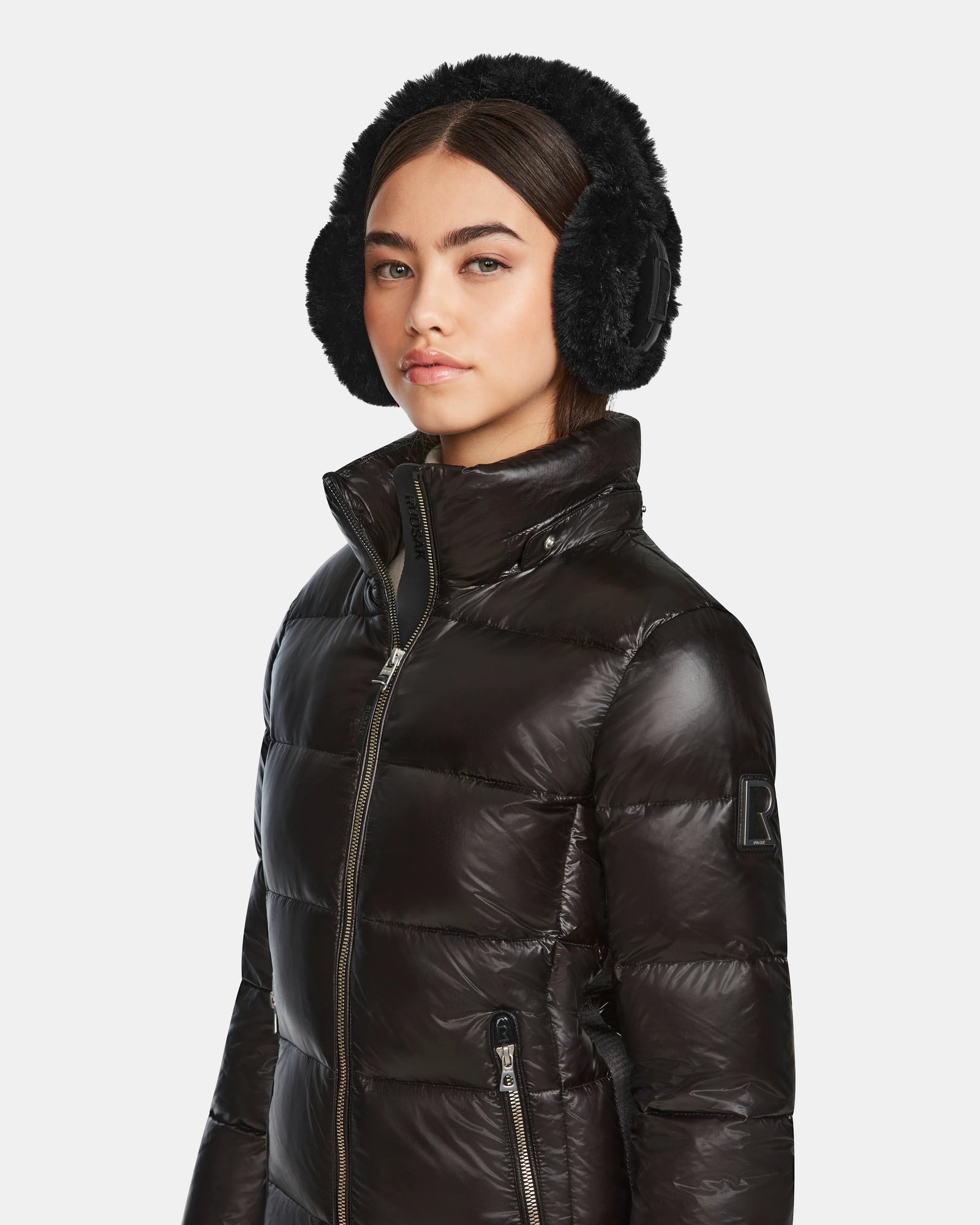 A woman wears the Rudsak MIRRY S - L123545 Black winter coat and black furry earmuffs, standing against a plain light background with a neutral expression.