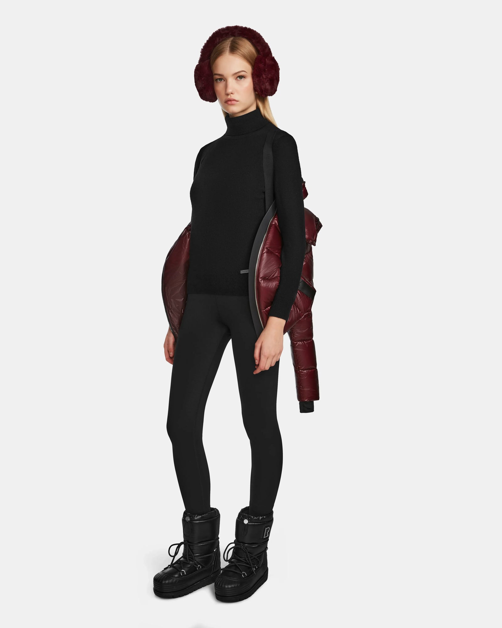 A woman faces forward against a plain white background, wearing black leggings, a black turtleneck, black snow boots, red earmuffs, and the Rudsak MIRRY S - L123545 Wine womens quilted down coat draped off her shoulders.