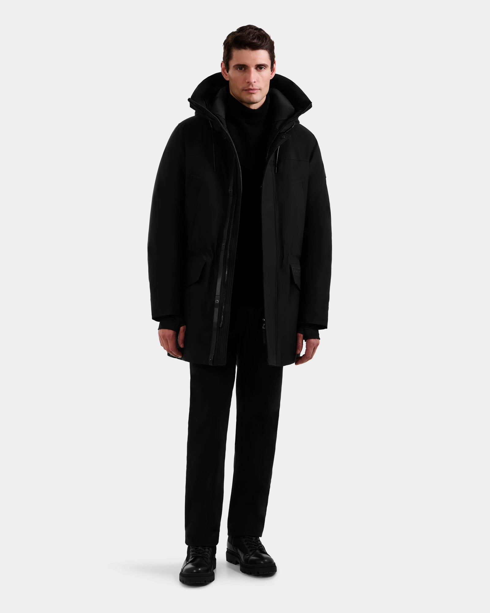 A man stands facing forward, wearing the Rudsak MITCHELL - M125513 Black men’s down parka with RDS-certified down, paired with black pants and shoes against a plain light gray background.