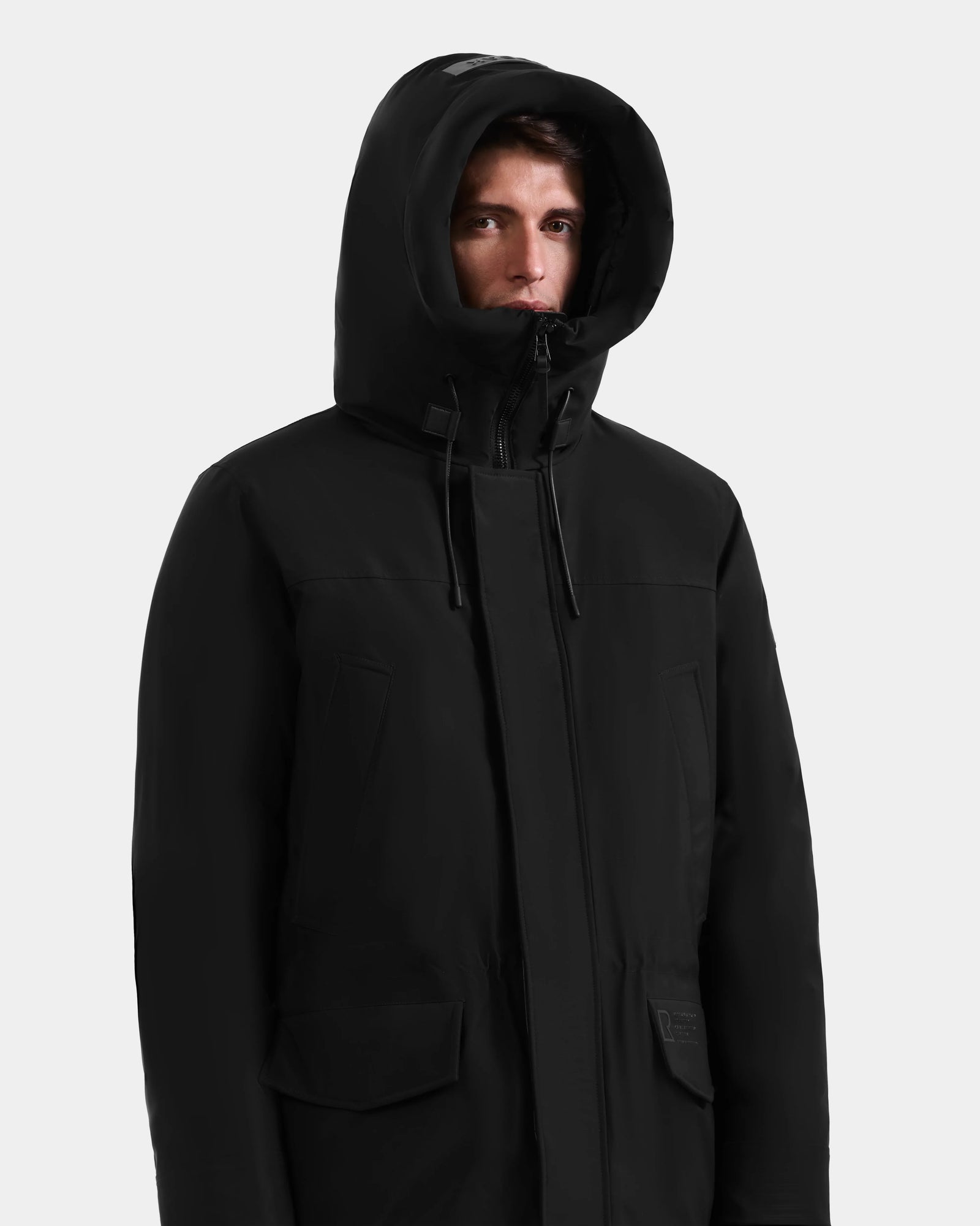 A person wearing the Rudsak MITCHELL - M125513 Black men’s down parka stands against a light gray background, looking slightly to the side with the hood up, partially covering their head and forehead.