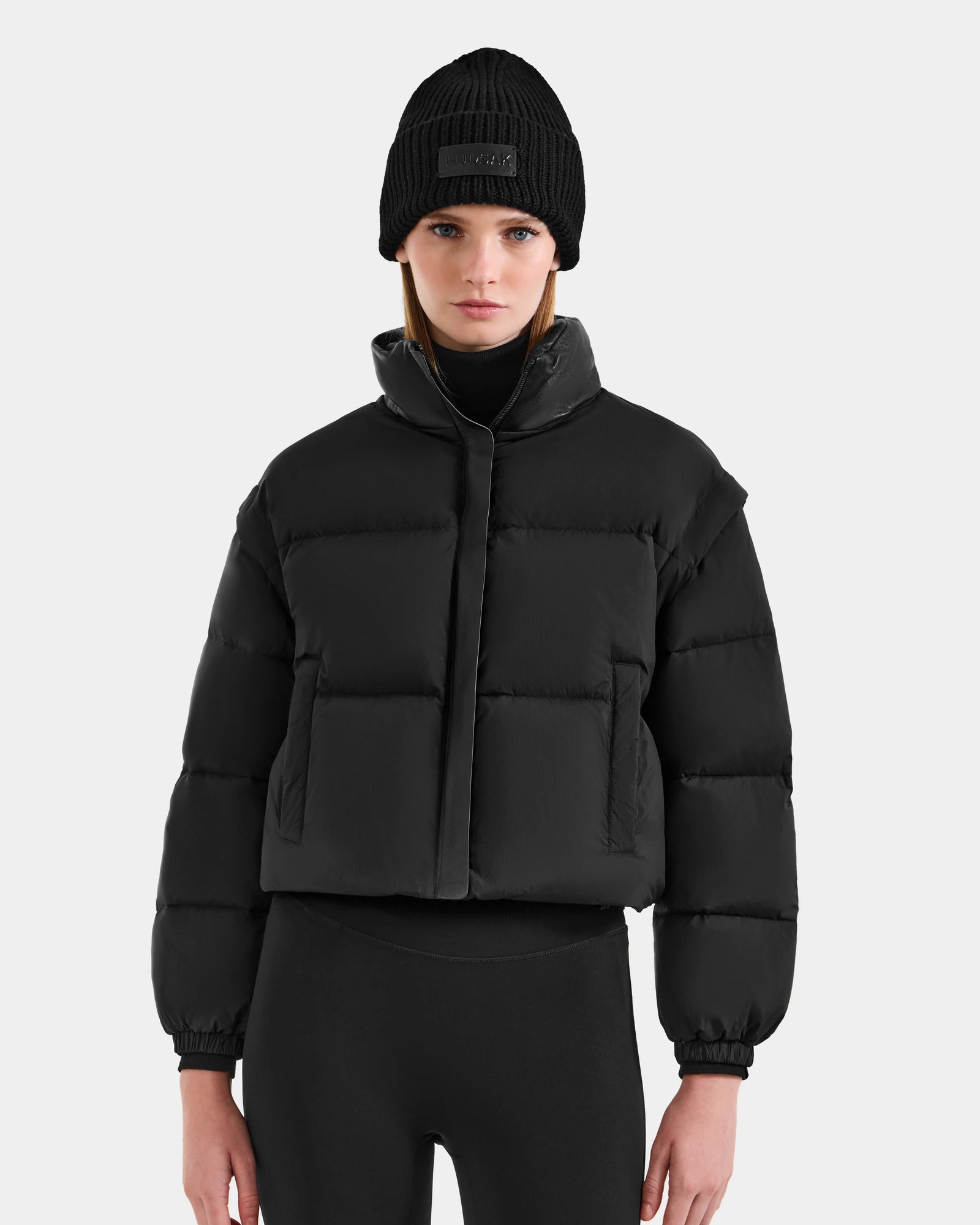 A woman stands against a light gray background, wearing the Rudsak OPRAH - L125546 Black women's cropped down puffer with removable sleeves, a black beanie, and leggings, looking directly at the camera with a neutral expression.