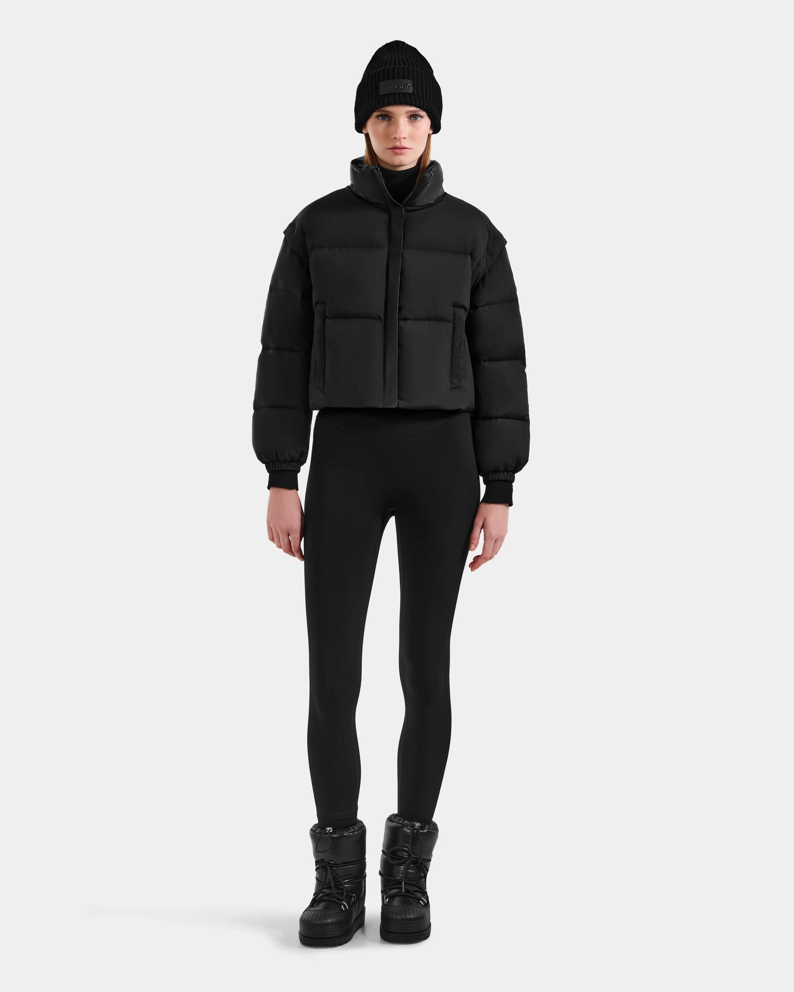 A person faces forward against a light gray background, wearing Rudsak's OPRAH - L125546 Black women's cropped down puffer with removable sleeves, paired with a black beanie, leggings, and boots.
