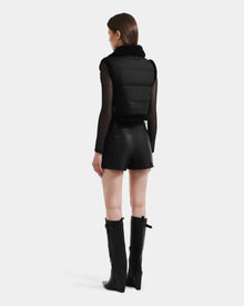 A woman faces away, wearing Rudsak's PAISLEY - L125516 Black vegan leather vest over a sheer top, paired with black leather shorts and knee-high black boots, set against a plain white background.