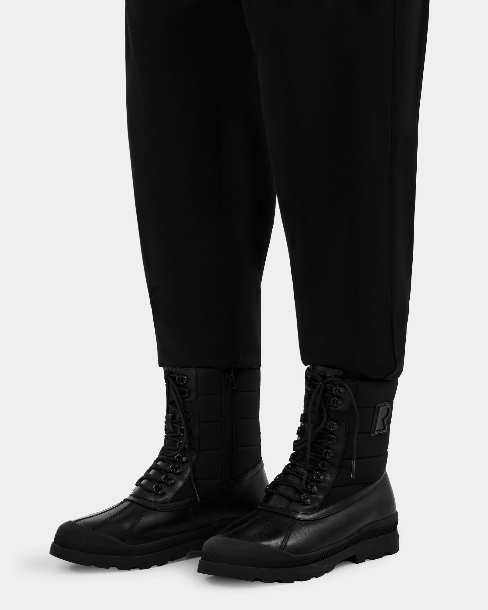 A person wearing black REAGAN - 6224501 boots by Rudsak stands against a plain, light-colored background.