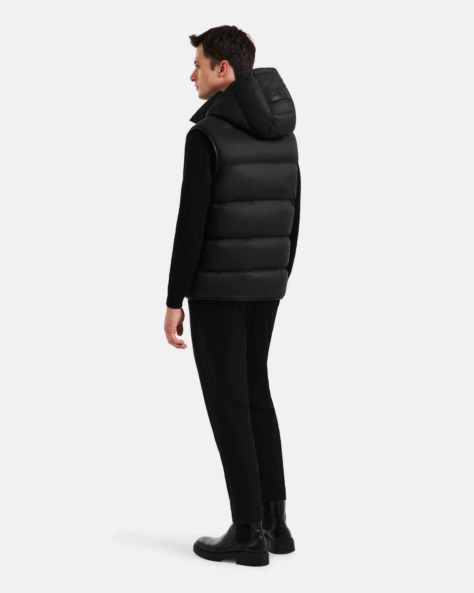A man faces away, wearing the Rudsak RICHARDSON - M125527 Black men’s hooded vest for water-repellent, lightweight warmth, styled with a black long-sleeve shirt, pants, and shoes against a plain white background.