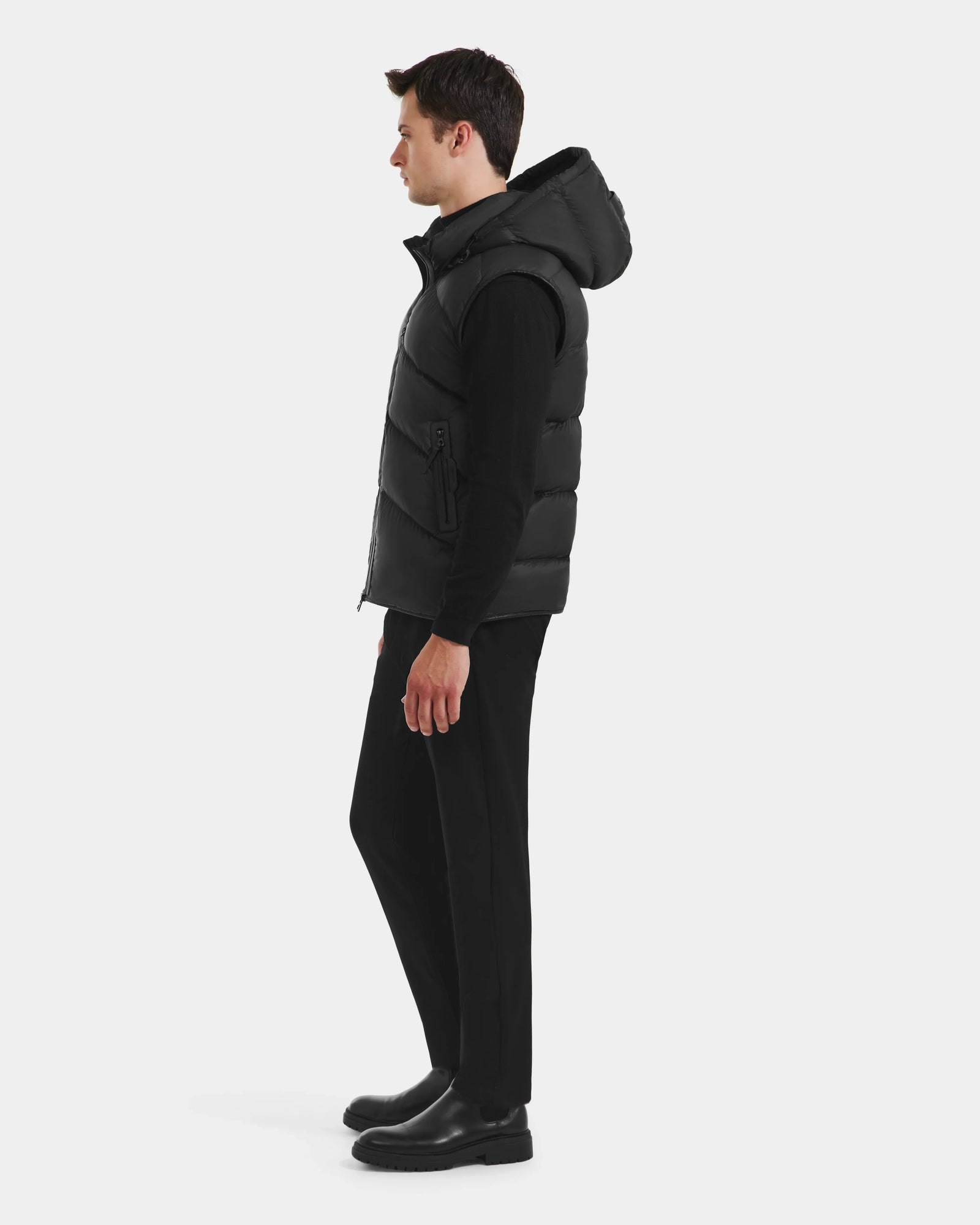 A man stands in profile against a white background, wearing the Rudsak RICHARDSON - M125527 Black men’s hooded vest for lightweight warmth, paired with a black long-sleeve shirt, black pants, and black boots.