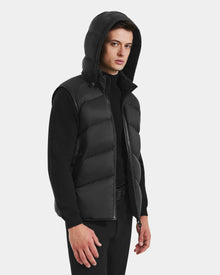 A man models the Rudsak RICHARDSON - M125527 Black men’s hooded vest, layered over a black sweater and pants, standing against a light background and highlighting its lightweight, water-repellent style.