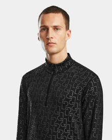 A man with short brown hair wears the Rudsak ROMEO - 6524525 Black men’s stretch active top, featuring a black high-collar mock neck zip-up with repeating light gray DG letters. The background is plain light gray.