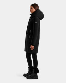 A person wears the Rudsak SARINA - L125550 Black women's down parka with RDS-certified down, high collar, patch pockets, black leggings, and winter boots, standing in profile against a light gray background.