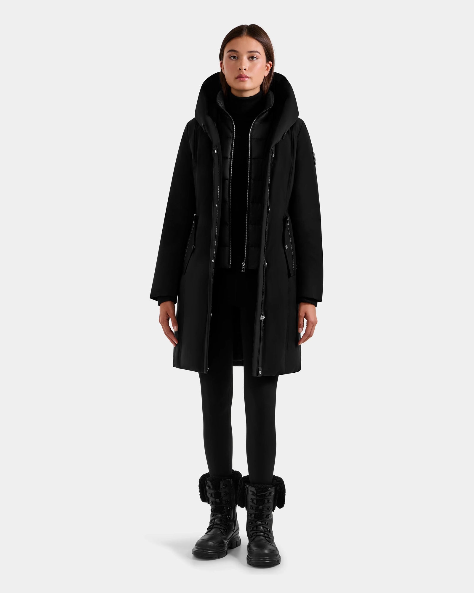 A woman faces forward wearing the Rudsak SARINA - L125550 Black semi-fitted parka with a high collar, paired with black leggings and black lace-up boots, posed against a plain light gray background.