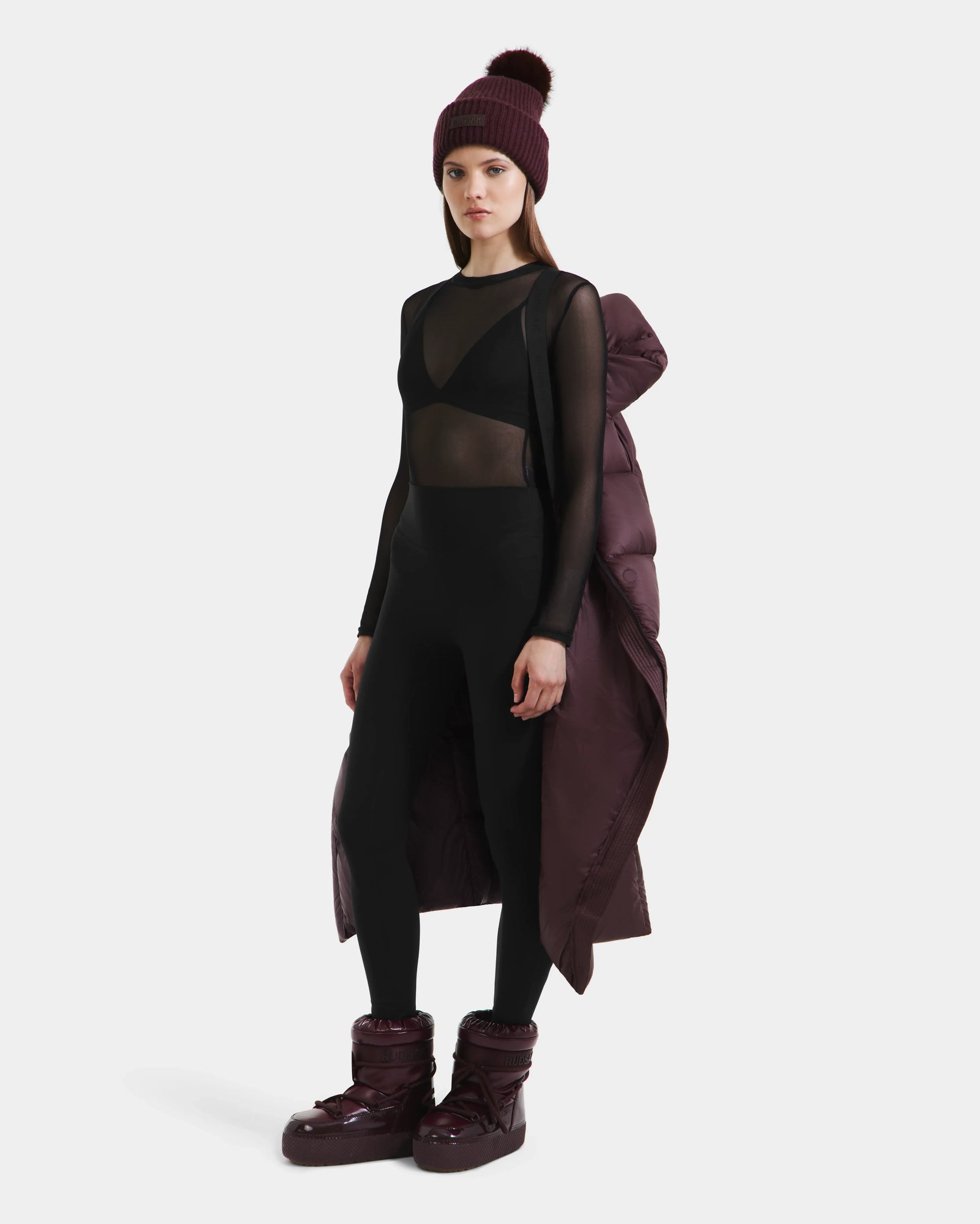 A woman in a sheer black bodysuit, black leggings, and maroon Rudsak SASHA - 8125686 Bordeaux accessories—a knit beanie, waterproof winter coat over her shoulders, and puffy boots—stands facing forward against a plain white background.