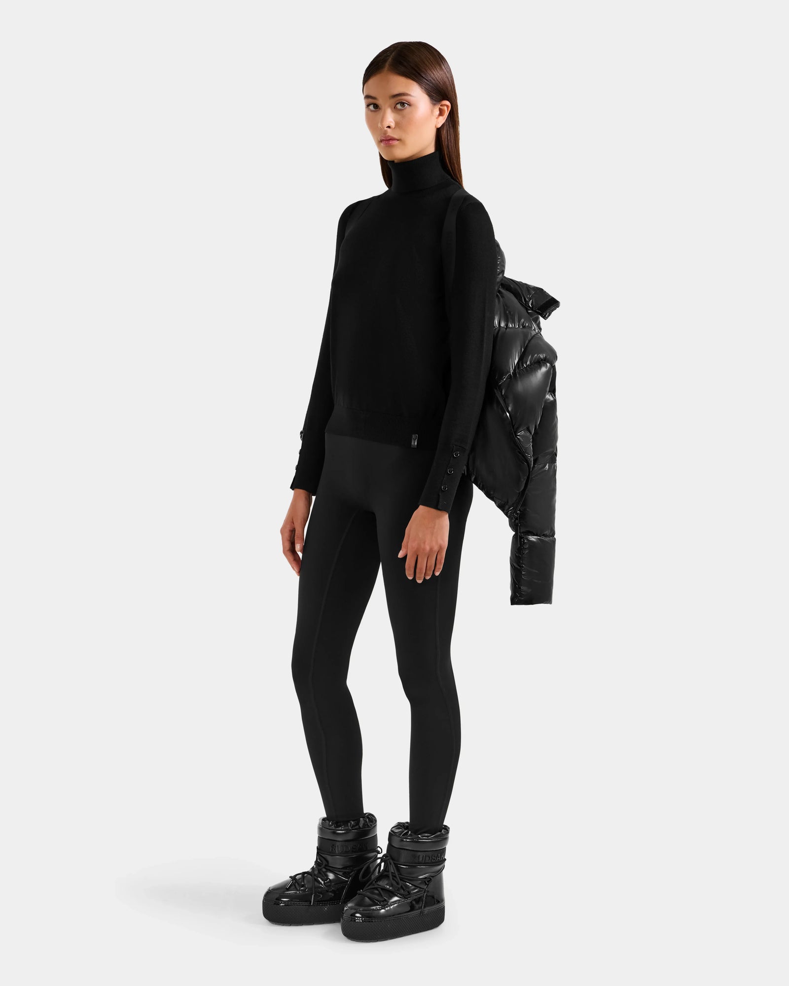 A woman stands against a plain background in Rudsak's STEFANIE S - L125585 Black short puffer, draped over one shoulder, paired with a black turtleneck sweater, leggings, and puffer boots.