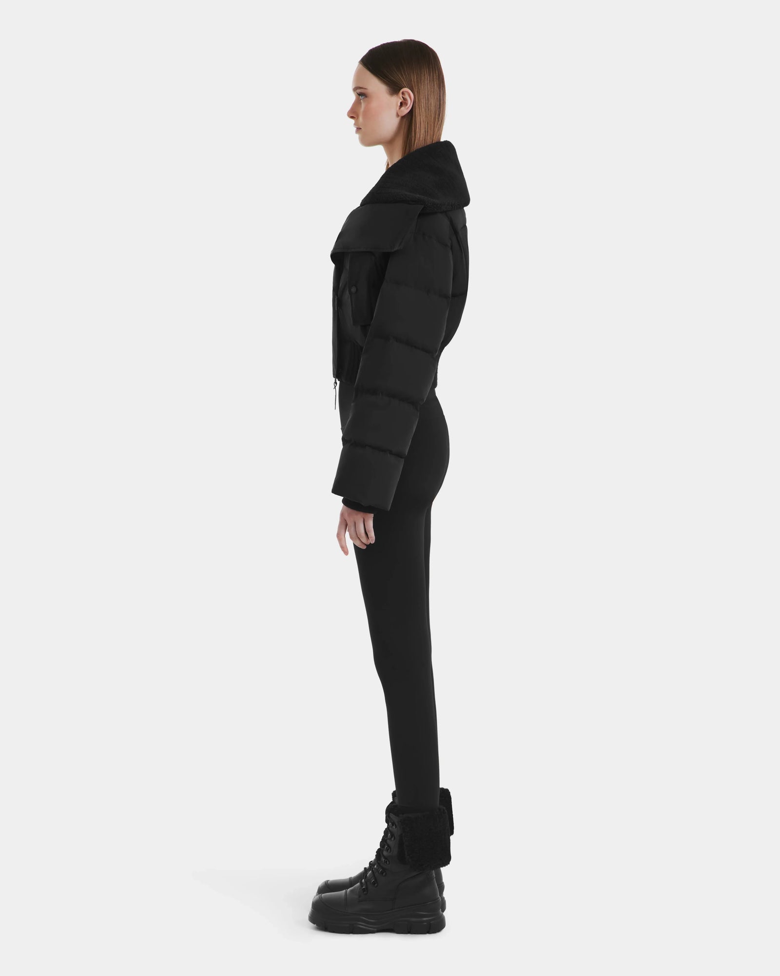 A woman stands in profile against a plain background, wearing the Rudsak YVETTE - L125557 Black women’s cropped down jacket with an oversized collar, paired with black leggings and black lace-up boots with fur trim.