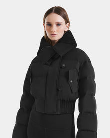 A woman with straight brown hair wears the Rudsak YVETTE - L125557 Black women’s cropped down jacket with an oversized collar and large front pocket, standing against a plain white background.