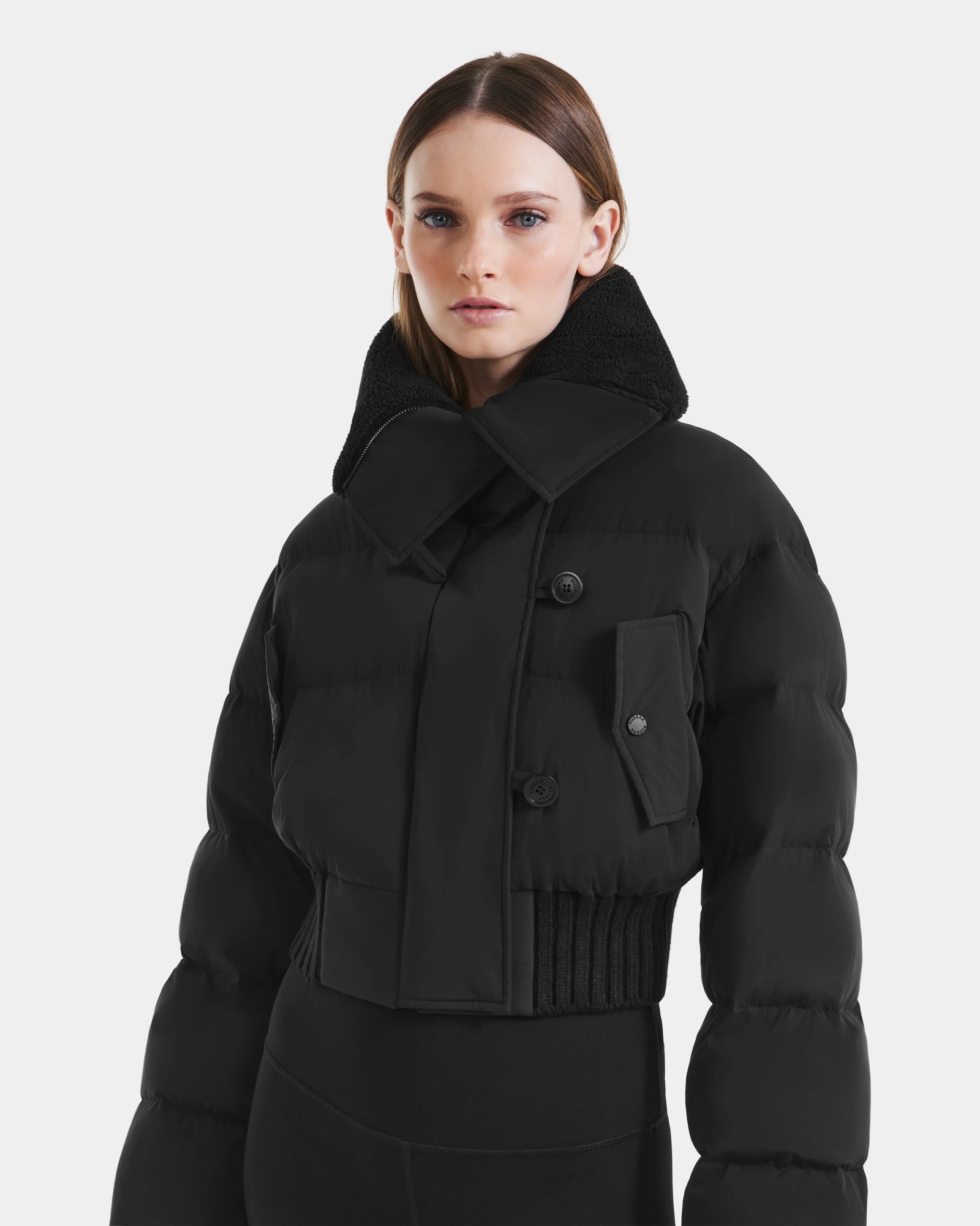 A woman with straight brown hair wears the Rudsak YVETTE - L125557 Black women’s cropped down jacket with an oversized collar and large front pocket, standing against a plain white background.