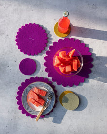 A table setting featuring graf lantz bright purple and yellow felt placemats, Merino wool trivet for heat protection, coasters, and tableware with watermelon slices and a pink drink on a light gray surface in natural sunlight.