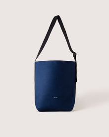A minimalist navy blue shoulder bag from graf lantz, featuring a black strap and crafted from sustainable materials, stands out against a neutral background with a small white logo near the bottom.