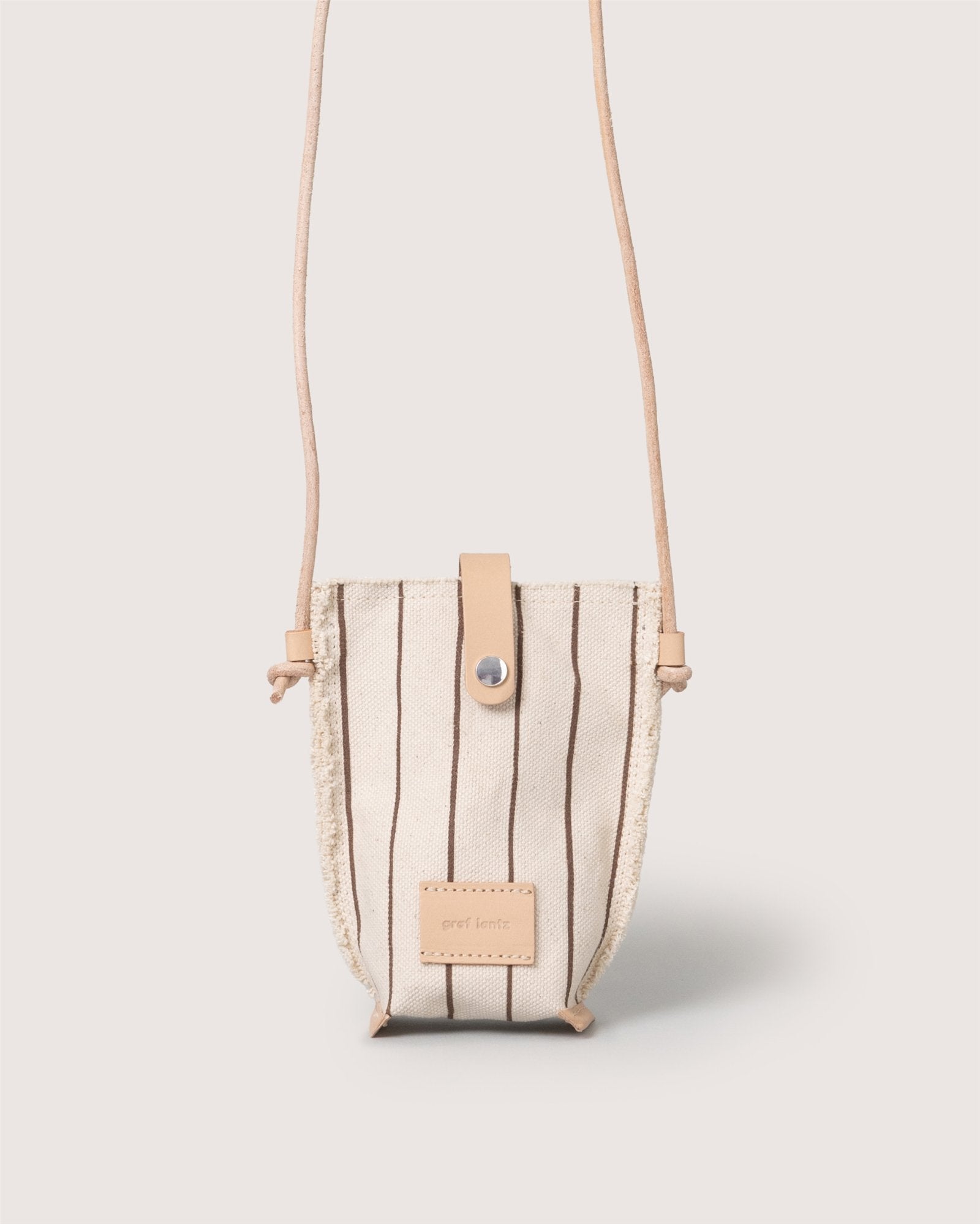 A compact, beige Graf Lantz Hana Canvas Phone Crossbody with vertical brown stripes, tan leather strap and snap closure, plus a front tan leather patch—shown against a light background for stylish hands-free convenience.