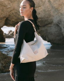 A woman stands on a beach with a Vachetta leather Hana Hobo bag from Graf Lantz. She wears a long-sleeve black dress and has her hair elegantly tied back, set against a serene backdrop of rocks and water, softly illuminated by the sunlight.