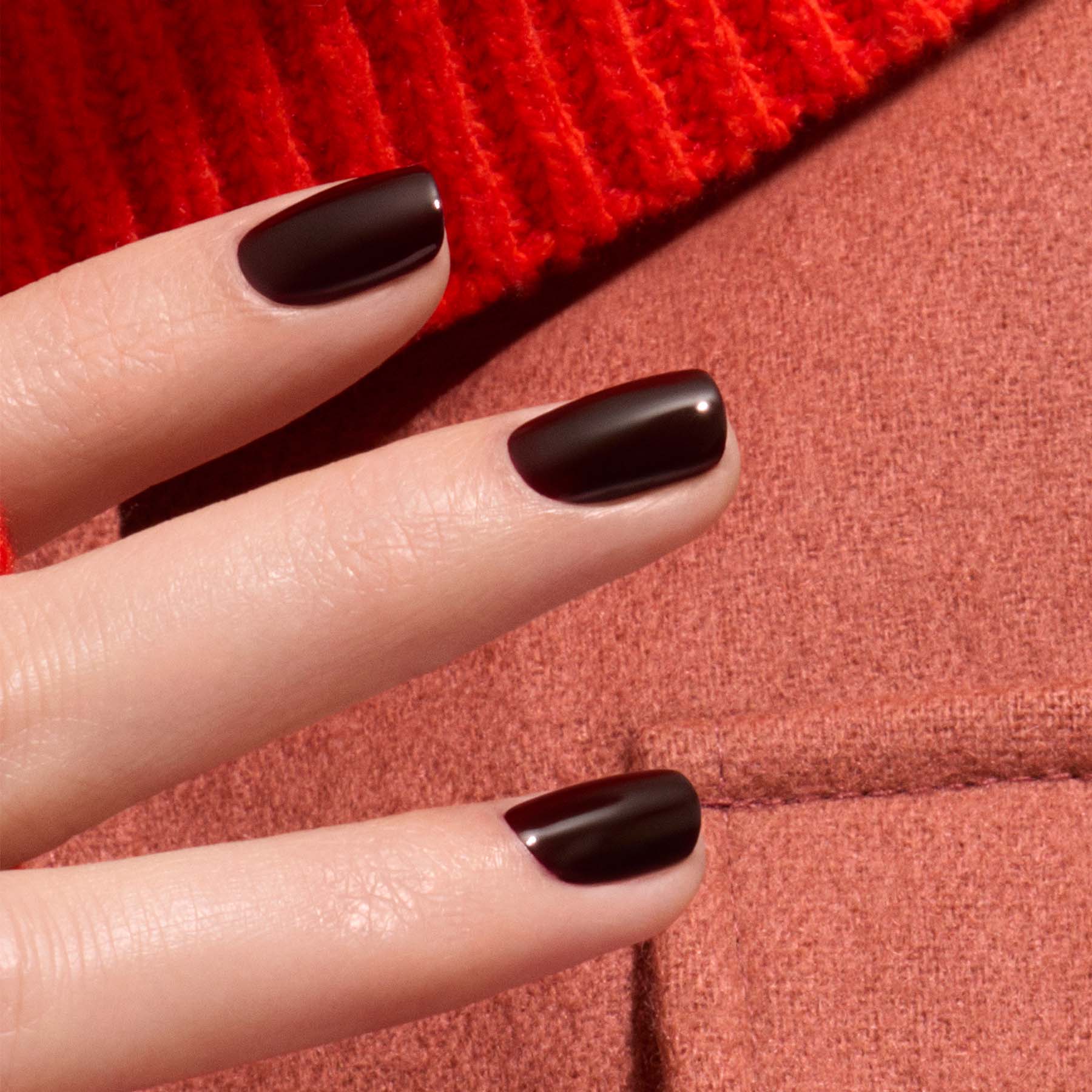 A close-up of a hand with neatly manicured nails painted in Manucurist Hollyhock, a rich black red gel polish, resting on textured red and pink fabric.