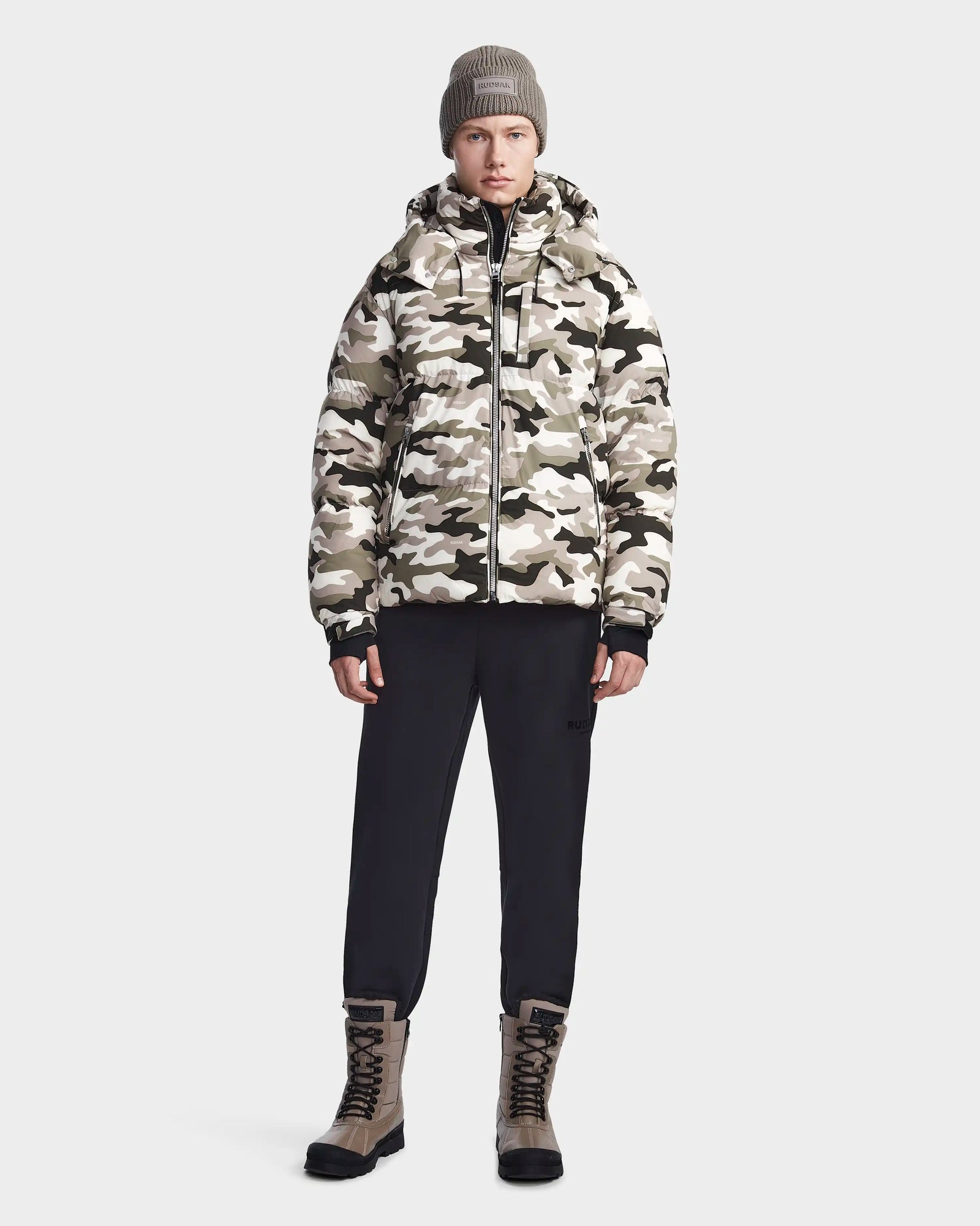 A person wearing a beige beanie, black pants, beige lace-up boots, and the Rudsak VINCENT M - 6123510 Camouflage men's down puffer jacket stands facing forward against a plain light gray background.